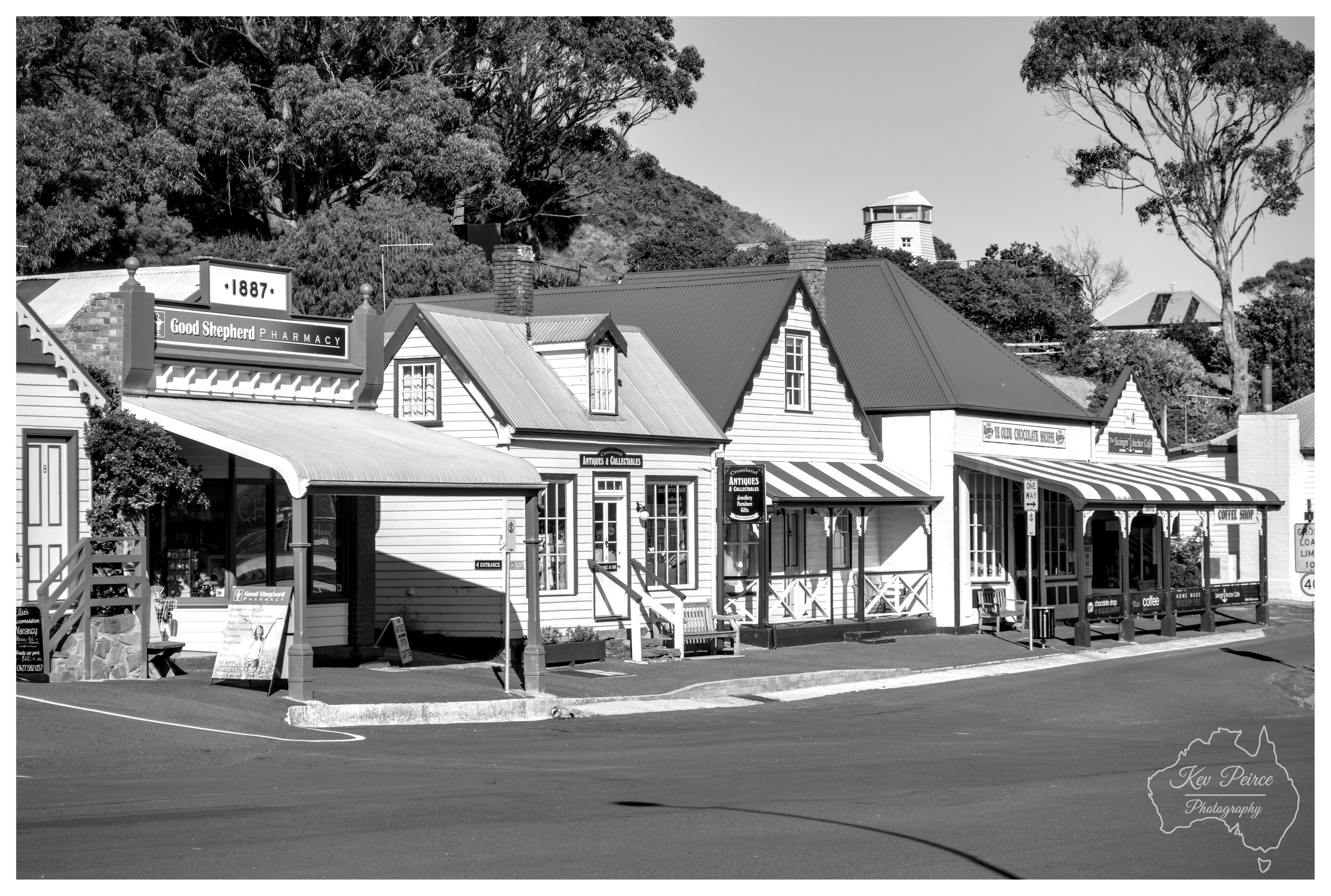 A black and white architectural shot showing a row of historic timber weatherboard shopfronts on a quiet town street. The buildings feature pitched corrugated roofs, decorative timber gables, and striped awnings, including the Good Shepherd Pharmacy dating back to 1887. The wide, empty asphalt roadway curves through the foreground under clear daylight, with eucalyptus trees and a hill rising behind the rooftops.