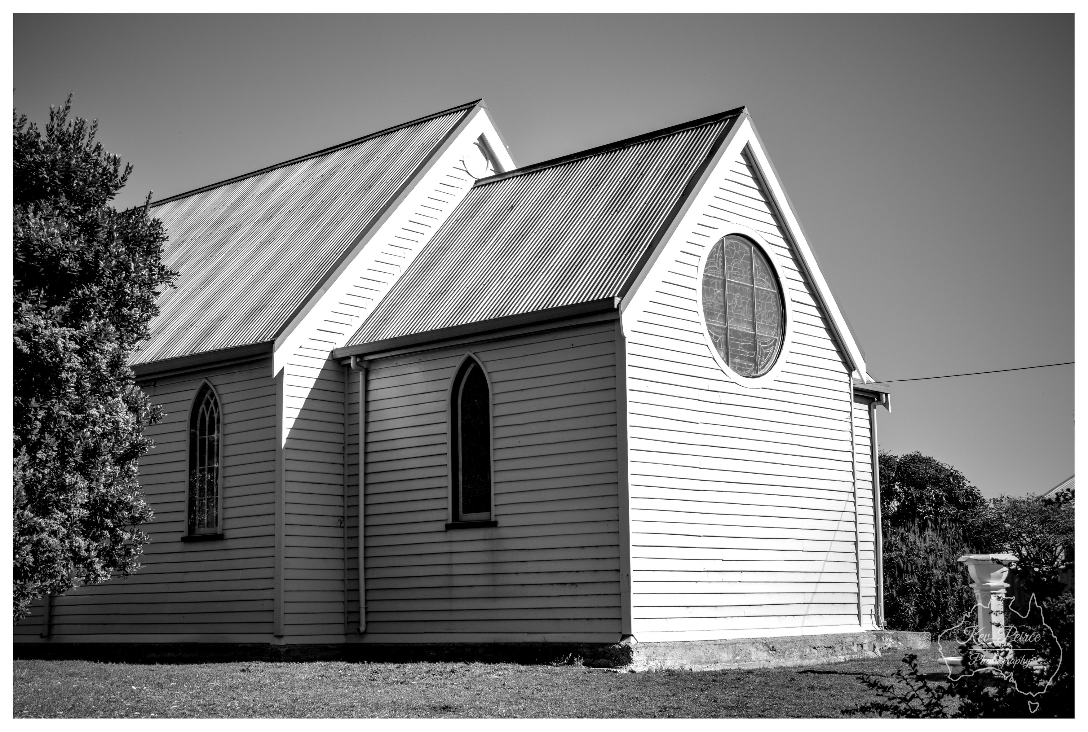 A black and white architectural shot showing the rear and side profile of a historic weatherboard church. The structure showcases steeply pitched corrugated metal gables, narrow pointed Gothic lancet windows, and a large circular lead light window on the main gable wall. Deep shadows highlight the horizontal timber weatherboards under bright daylight.