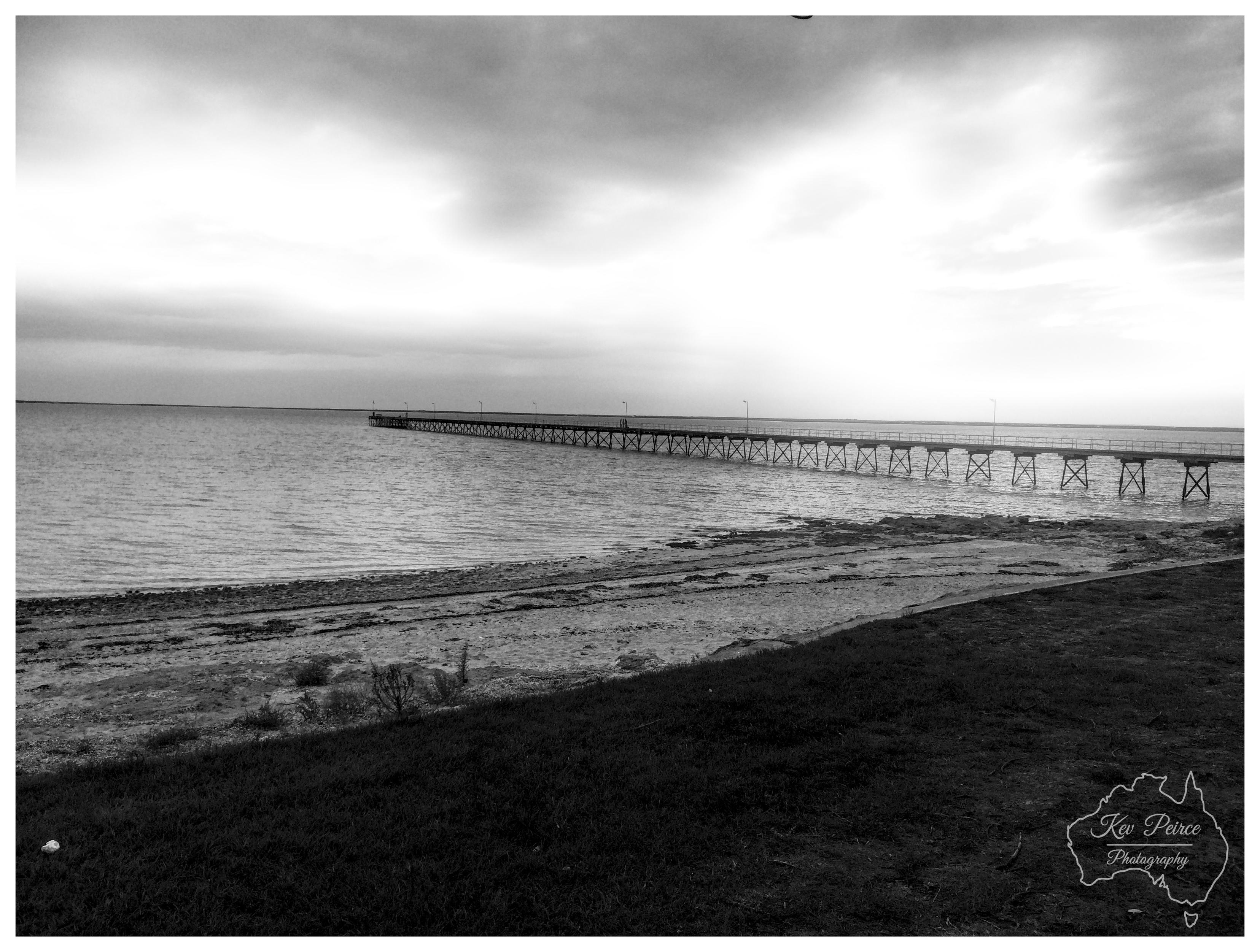 A monochrome landscape photograph capturing a long coastal jetty stretching into calm water under an overcast sky. The foreground shows a grassy, dark shoreline sloping down towards a sandy, pebble strewn beach. In the mid ground, the narrow wooden and steel pier extends horizontally towards the left across the calm sea. The horizon is low and flat, beneath heavy, textured grey clouds that fill the upper portion of the frame with soft, diffused light. A subtle vignette darkens the corners, and a circular outline watermark is visible in the lower right corner.