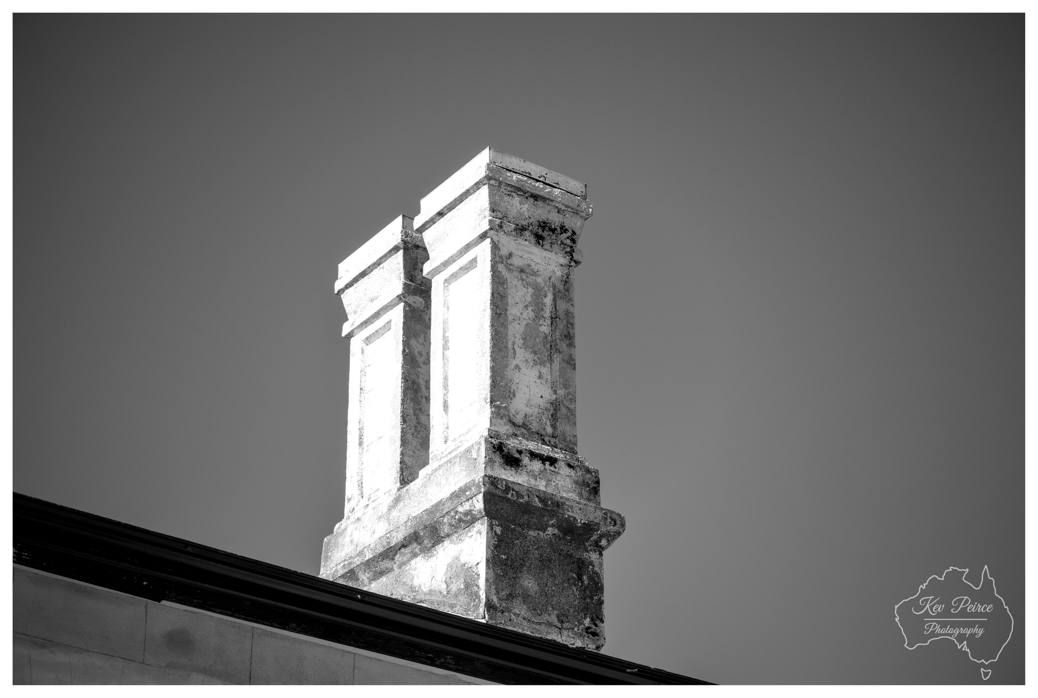 A black and white, low angle architectural photograph focusing on ornate twin chimney stacks rising above a heritage roofline. Strong sunlight illuminates the pale, weathered textured render and moulded panels on the front facade, casting deep shadows along the sides. The background is a smooth, clear sky rendered in deep grey tones.