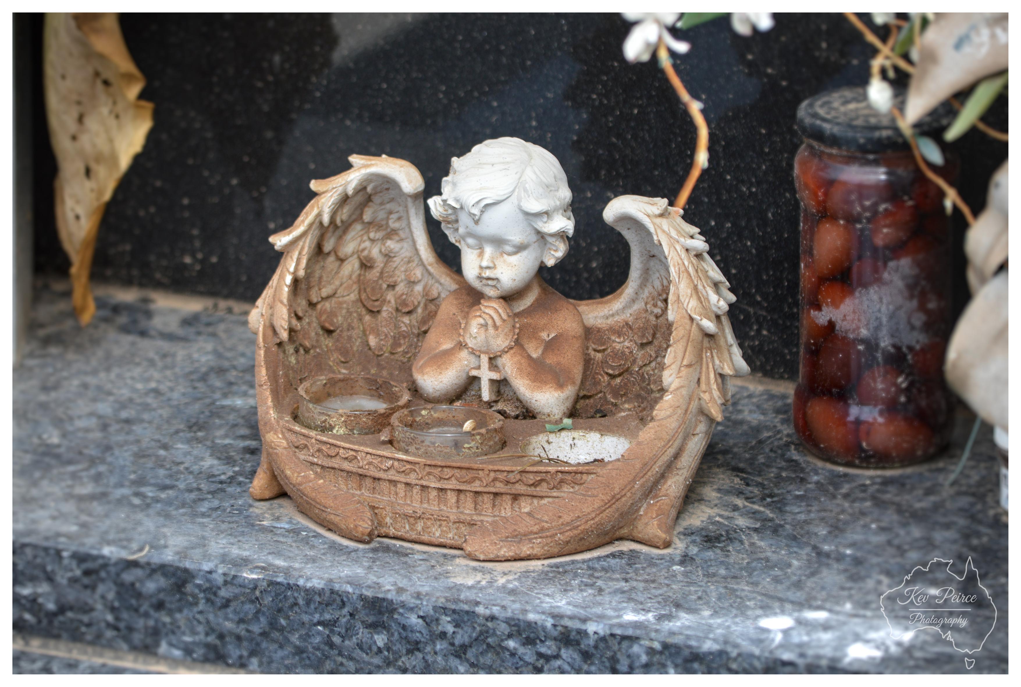 A close up, horizontal shot of a weathered stone figurine of a small angel with large wings, resting on a dark polished granite memorial surface. The angel has its hands clasped in prayer holding a small cross, and the base features small circular candle holders. To the right, a glass jar filled with preserved red objects sits beside a faint artificial flower stem. The bottom right corner displays a subtle watermark for Kev Peirce Photography.