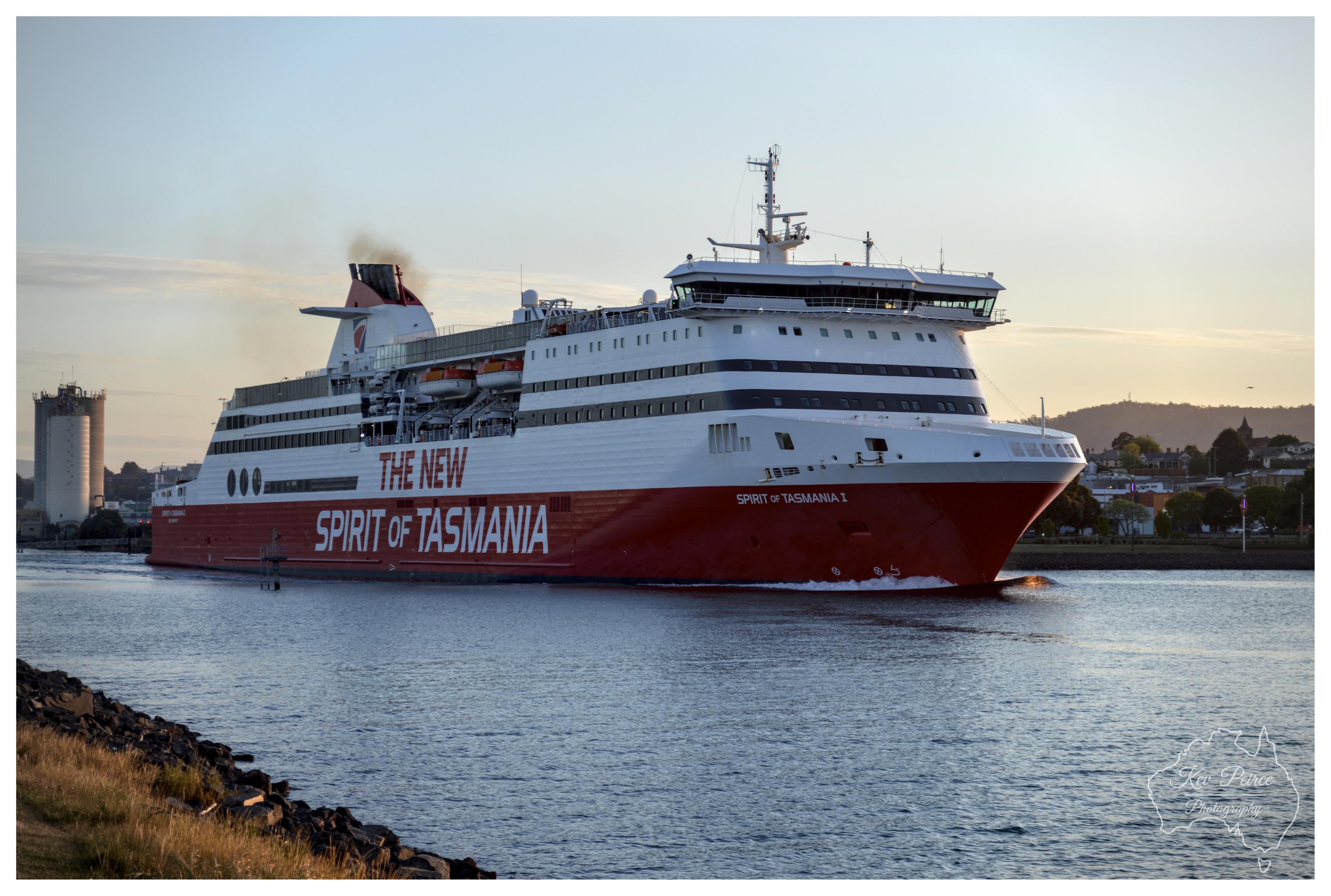 A horizontal medium shot capturing the large red and white Spirit of Tasmania ferry moving through a calm river channel. The ship features multiple decks, large windows, and bold text along the hull, illuminated by soft golden hour light from the side. A rocky shoreline lines the lower left foreground, and a subtle watermark for Kev Peirce Photography appears in the bottom right corner.