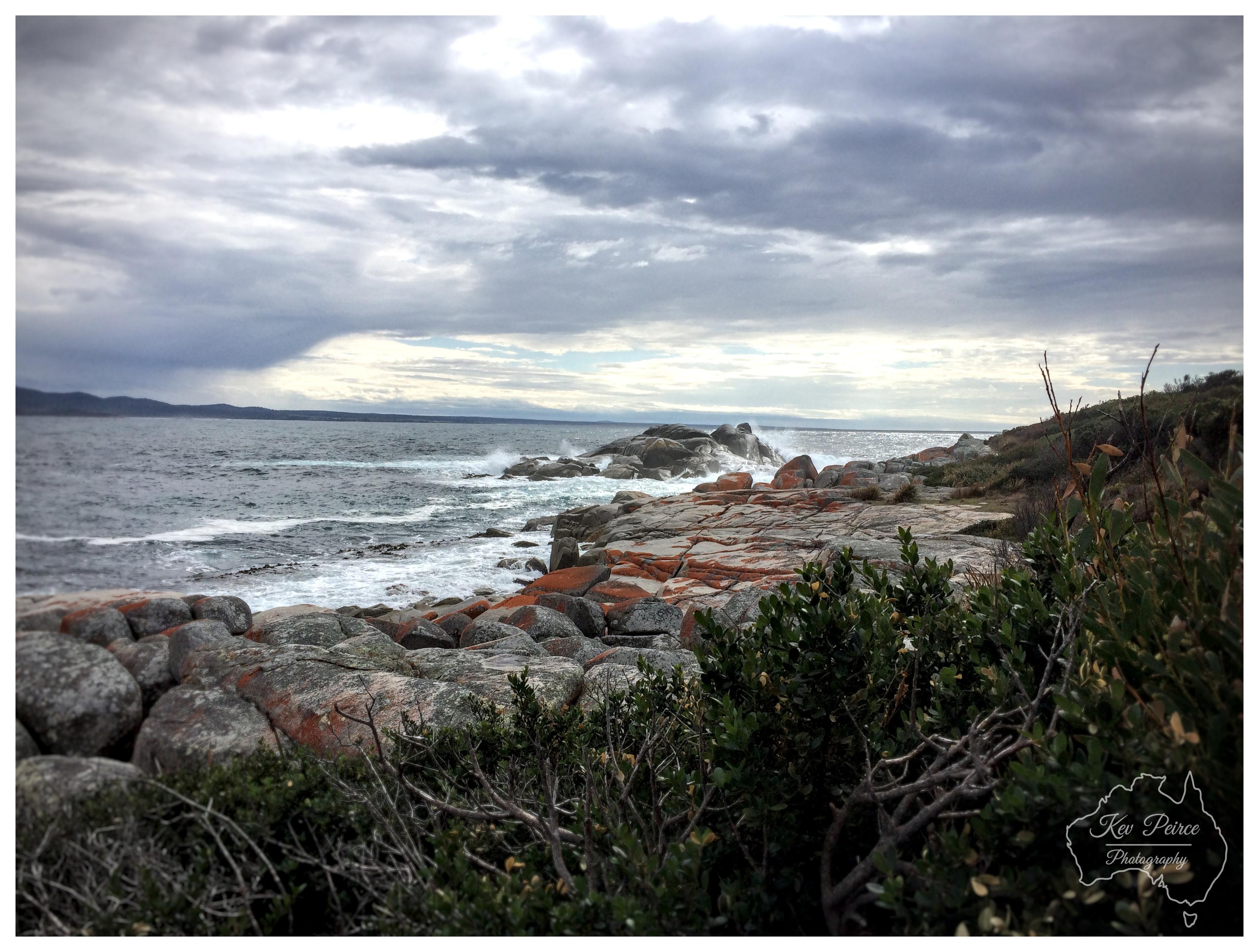 A wide coastal shot showcasing rough ocean waves crashing against distant rocky reefs under a dramatic, cloud-filled sky. Foreground bushes and coastal vegetation frame the scene on the right, looking out over weathered grey and orange granite rocks along the shoreline. A watermark for Kev Peirce Photography appears in the bottom right corner.