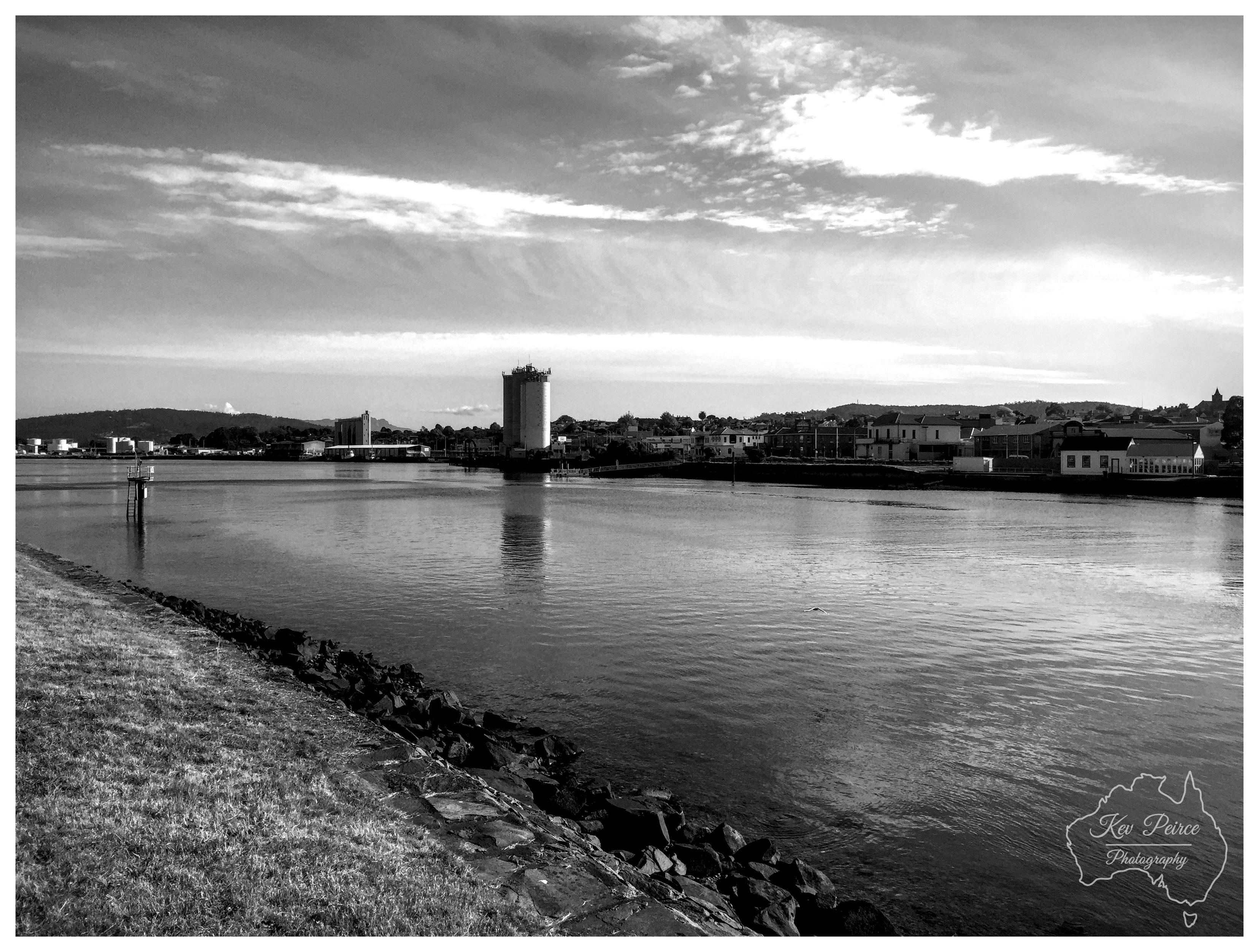 A horizontal black and white wide shot of a calm river flowing past a rocky embankment on the left. In the midground, the opposite bank features a town skyline dominated by a tall, cylindrical silo and low slung buildings under a dynamic, cloudy sky. A faint watermark for Kev Peirce Photography is visible in the bottom right corner.