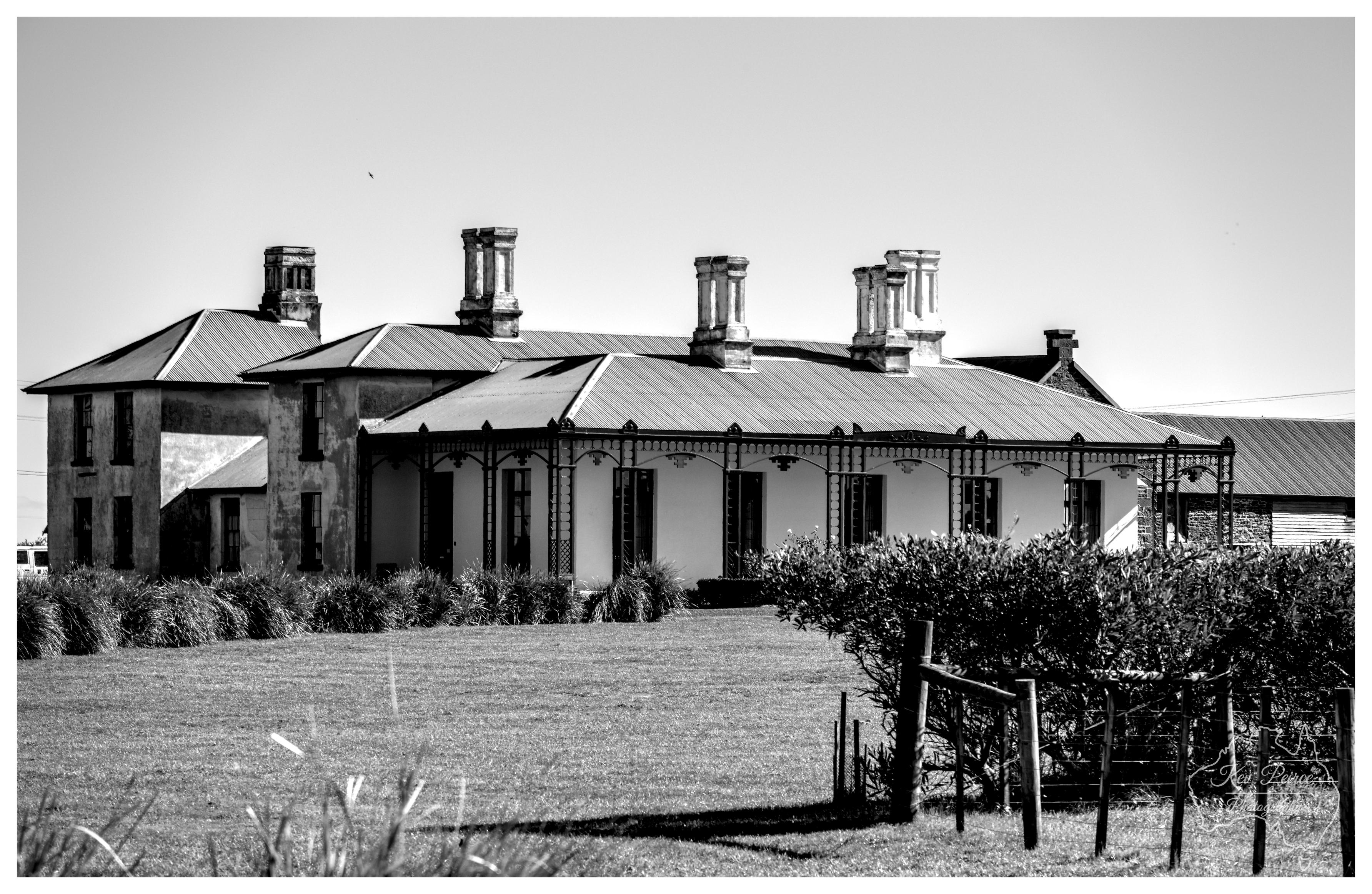A horizontal black and white medium shot of historic Highfield House, featuring a sprawling single storey homestead with a corrugated iron roof and multiple prominent chimneys. The veranda is supported by dark, slender posts, and the surrounding grounds include a neatly mown lawn, native grasses, and a wooden fence on the right. A watermark for Kev Peirce Photography sits in the lower right corner.