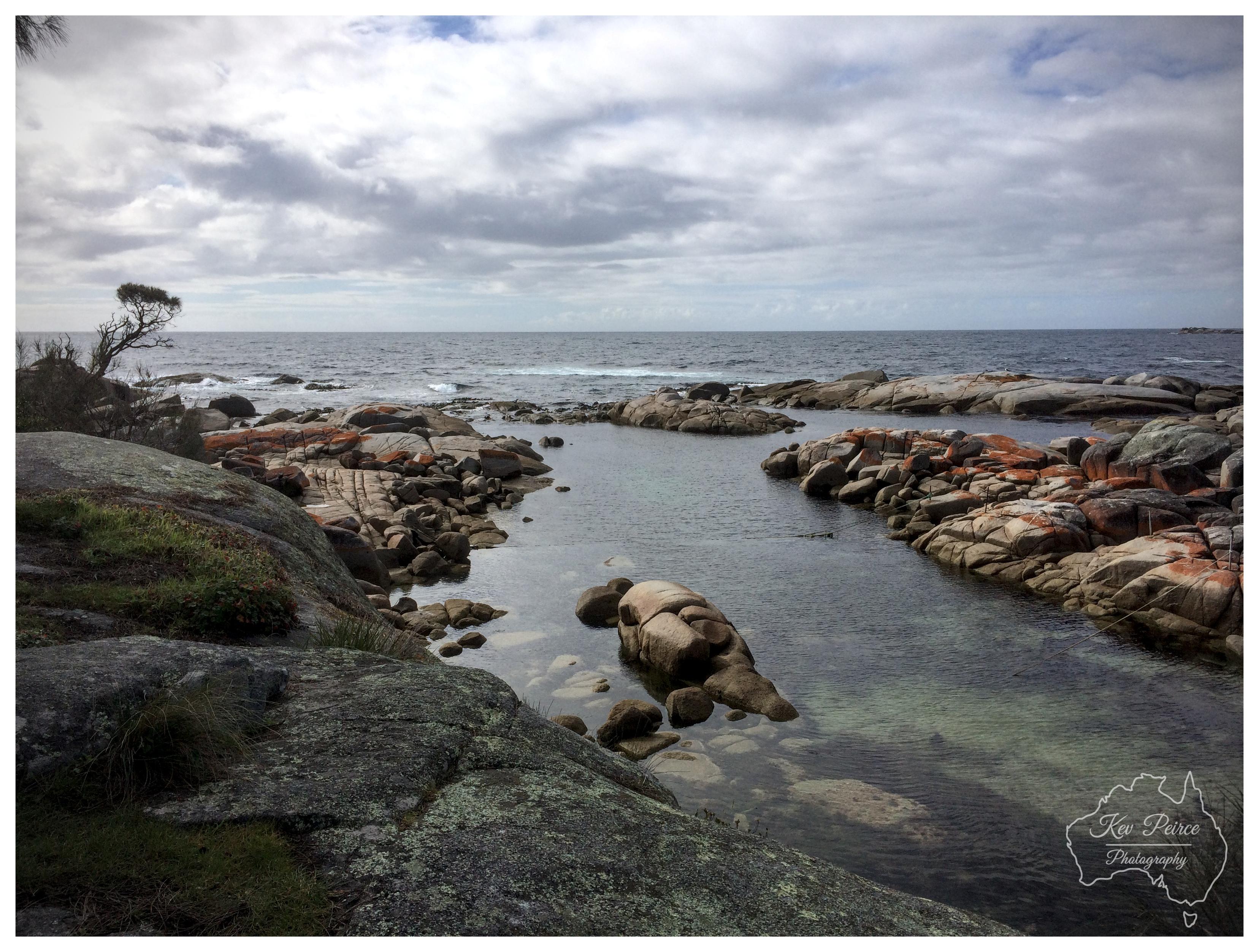 A horizontal wide shot of a rocky coastline in Tasmania, featuring smooth granite boulders stained with orange lichen. A calm channel of clear water winds through the rocks toward the open sea under a heavily clouded sky. A solitary windswept tree stands on the left headland. A subtle watermark for Kev Peirce Photography sits in the lower right corner.