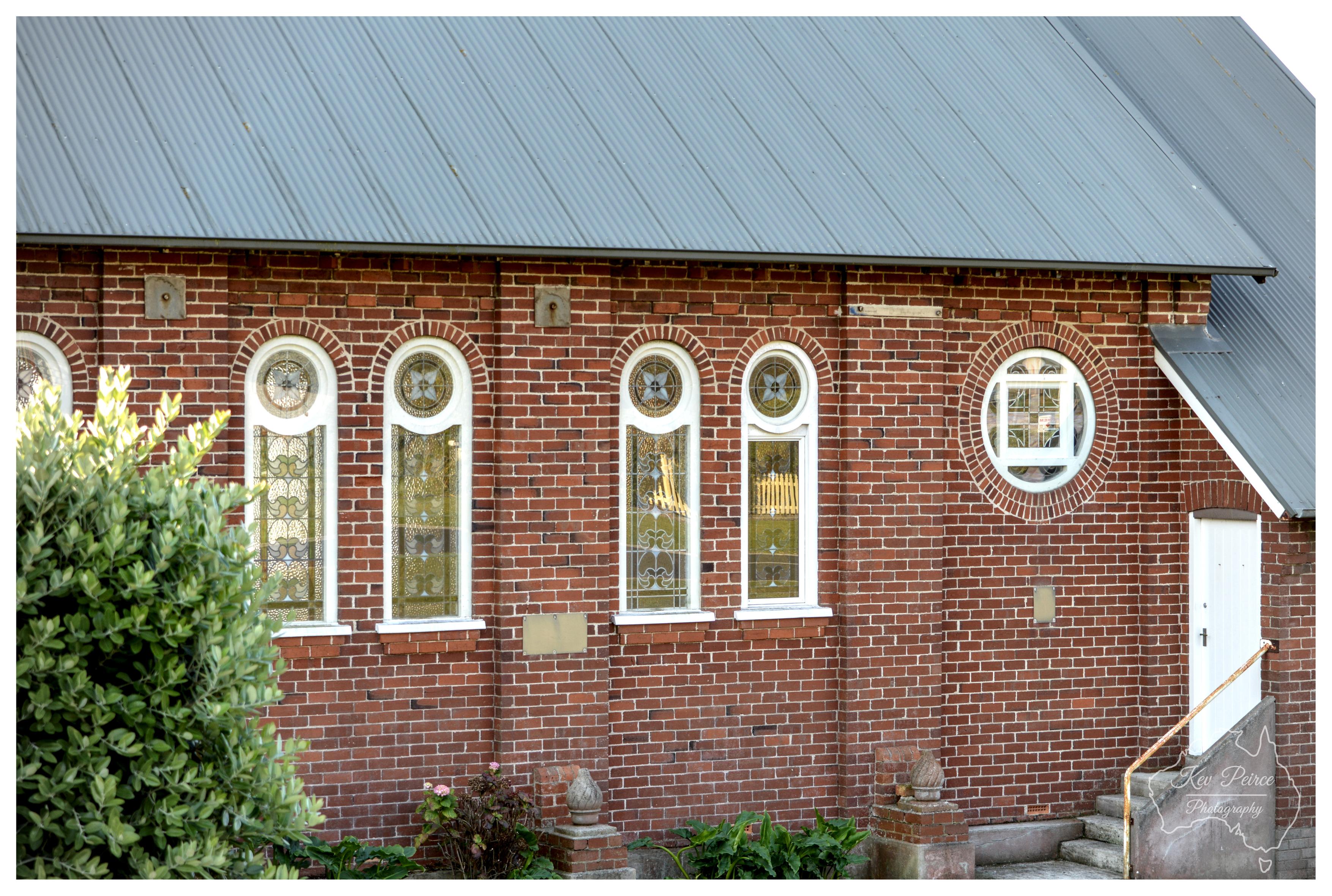 A close, straight on exterior shot of a red brick church wall illuminated by soft daylight. The facade features four tall, narrow arched windows fitted with decorative stained glass, alongside a small circular window framed with white masonry. A corrugated iron roof overhangs the top edge. Green foliage occupies the lower left corner, and a small flight of concrete steps leads to a closed white side door at the far right.