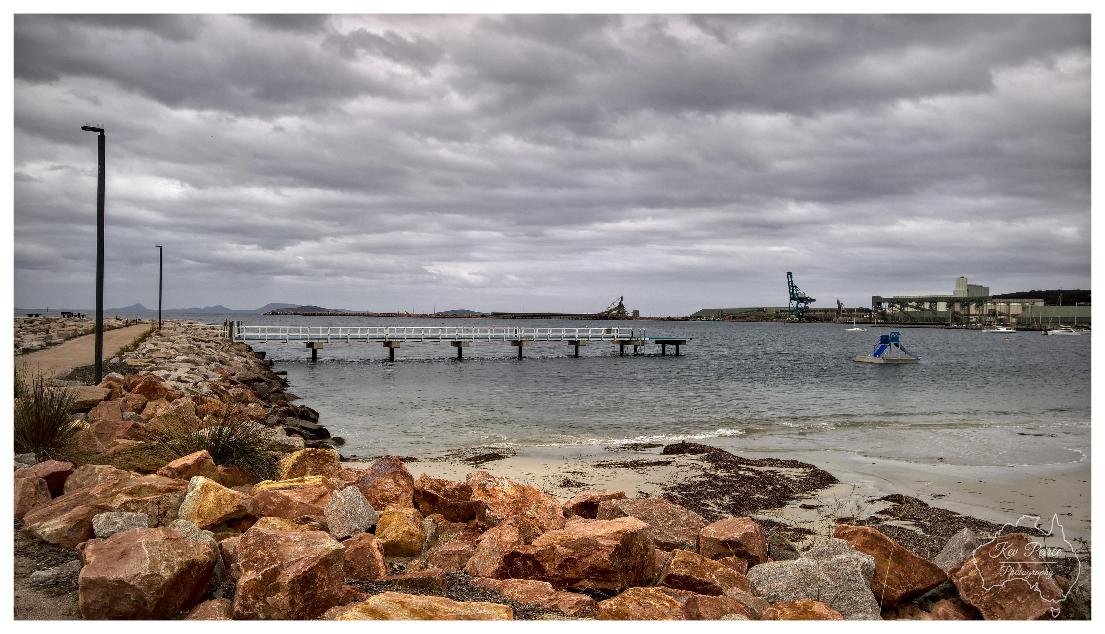 A horizontal wide shot of a coastal harbour under a heavy, overcast grey sky. In the foreground, warm reddish boulders line a sandy beach and a paved walkway. A white wooden jetty extends into the calm, dark blue water. In the distance, industrial port structures, cranes, and low hills line the horizon. A subtle watermark for Kev Peirce Photography appears in the bottom right corner.